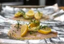 A close-up photograph of raw white fish fillets on aluminum foil, topped with lemon slices, herbs, and olive oil, prepared for baking.