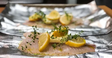 A close-up photograph of raw white fish fillets on aluminum foil, topped with lemon slices, herbs, and olive oil, prepared for baking.